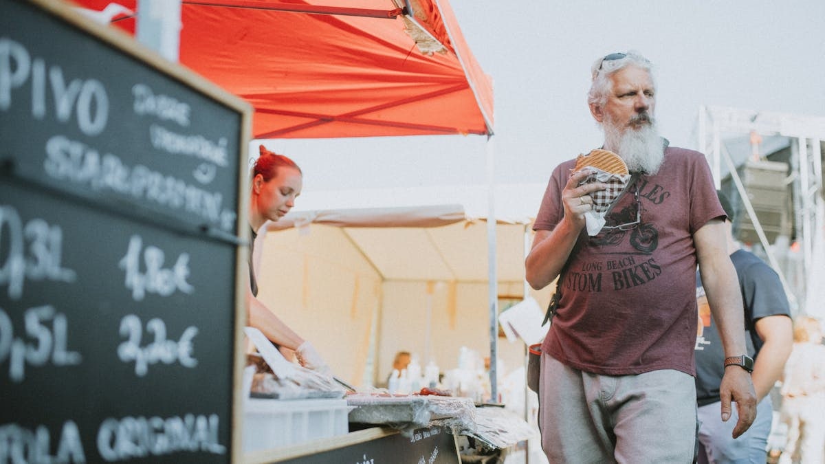 Man eating a burger at a street food market with beer menu in view, vibrant street scene.